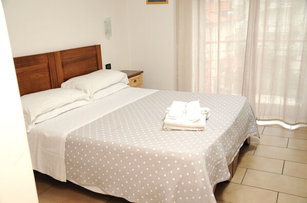 A neatly made bed with a polka dot bedspread and white pillows, a wooden headboard, a small bedside table, and a window with sheer curtains.