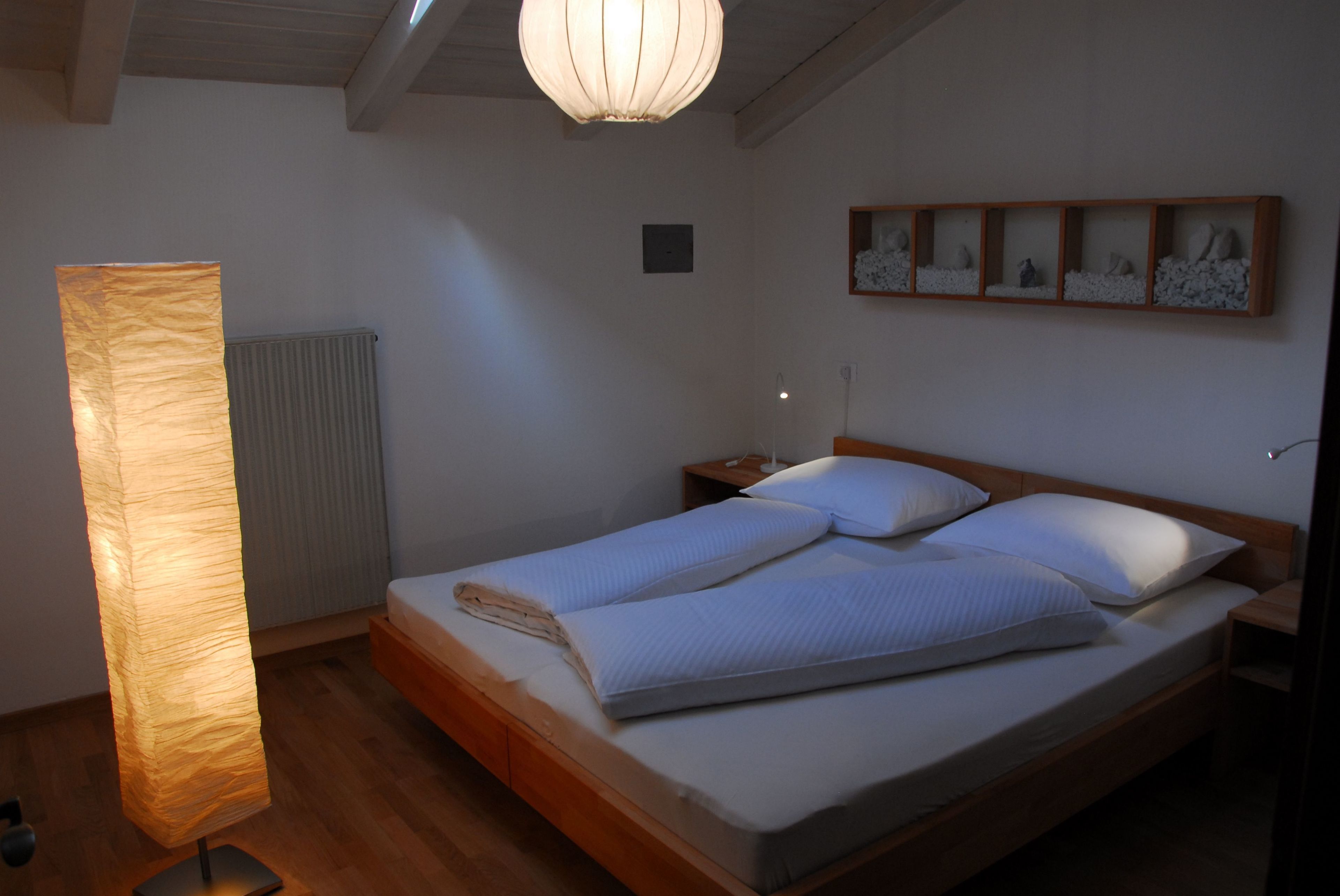 Ein Schlafzimmer mit einem hölzernen Bett, weißer Bettwäsche, einem Nachttisch, einer Wandablage mit Dekoration und einer Stehlampe.