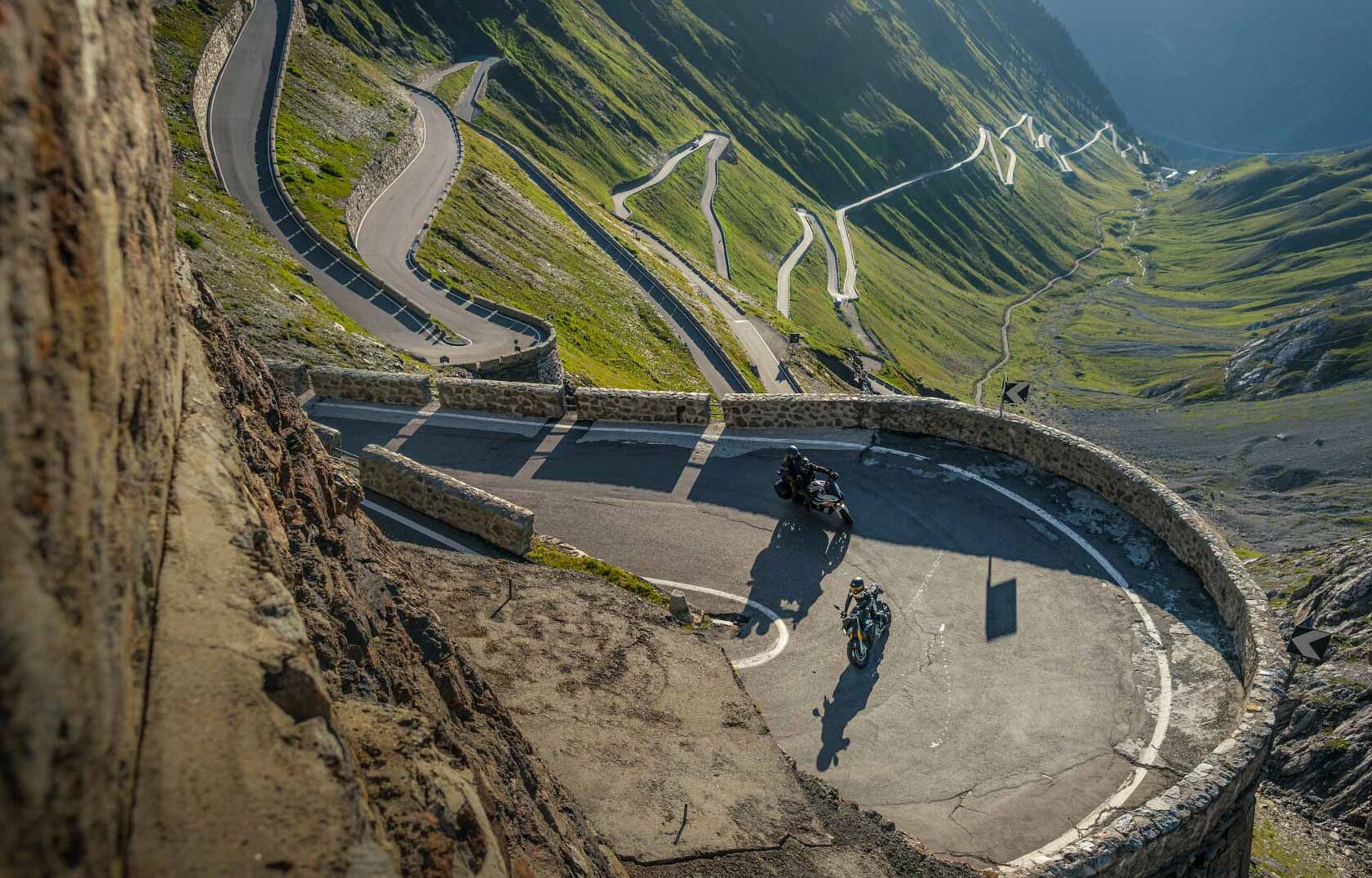 Una carretera sinuosa de montaña con barandilla de piedra, rodeada de colinas verdes y terreno rocoso.