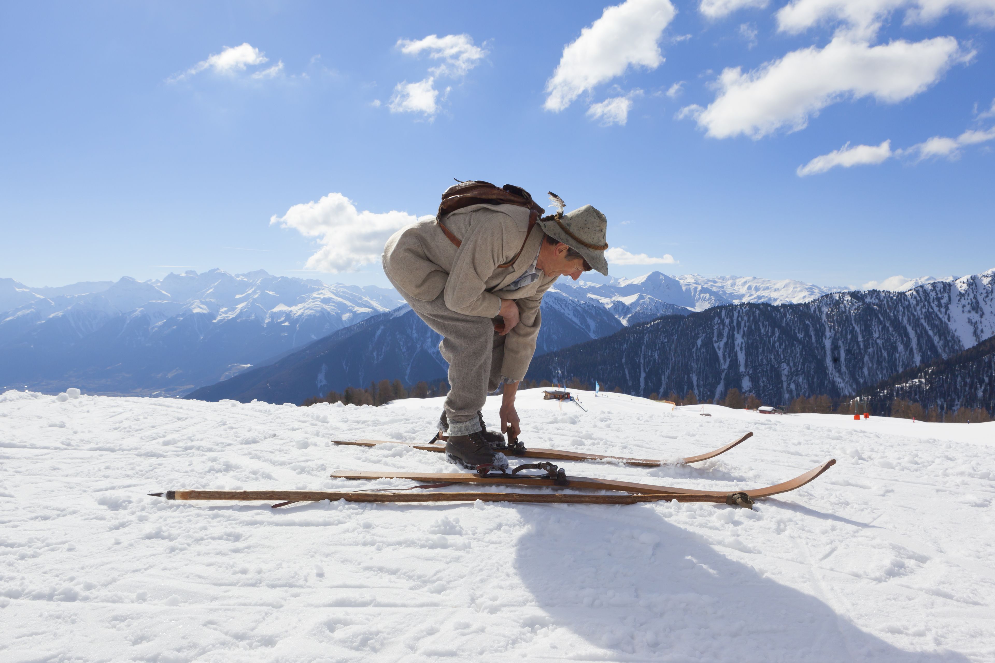 A person adjusting skis on a snowy slope with mountains in the background.