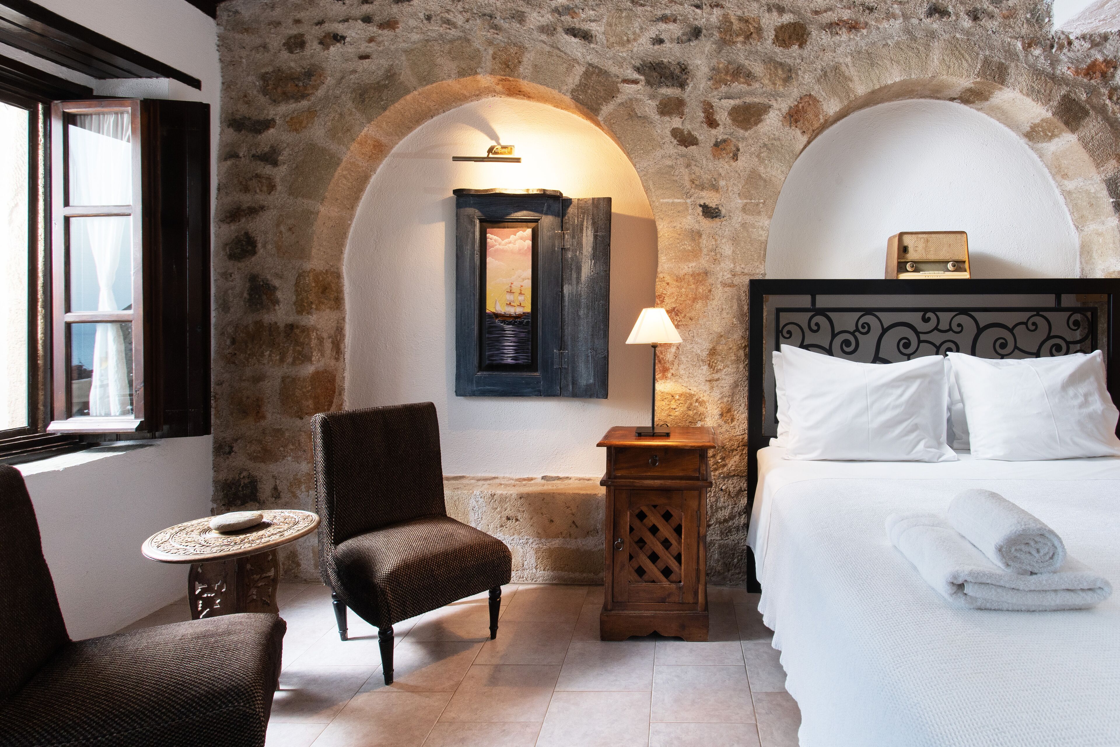 A room with a stone wall, a bed with white linens, a chair, a small table, and a lamp.