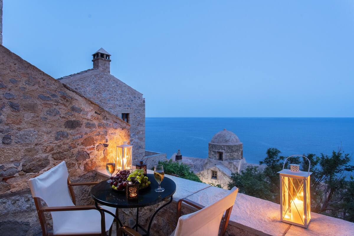 Un edificio de piedra con una mesa, sillas, un farol y vista al mar.