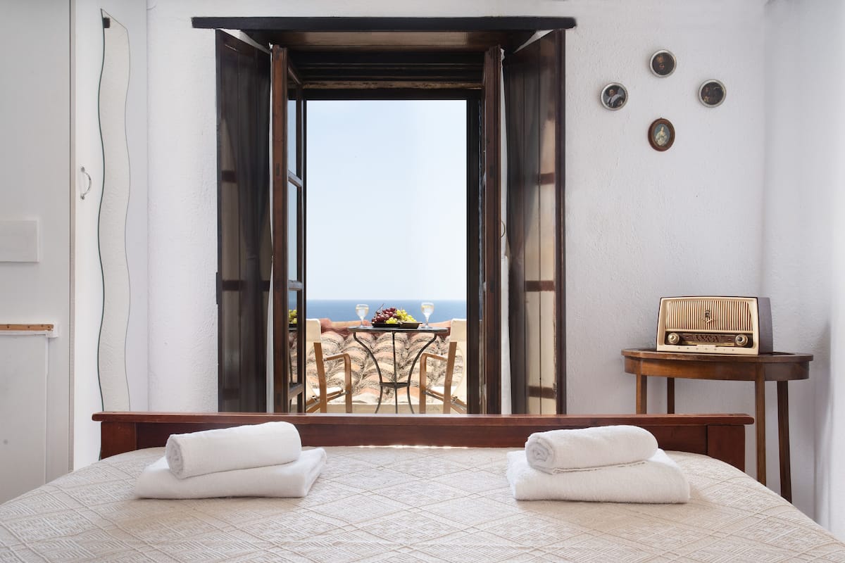 Habitación de hotel con cama, dos toallas, una mesa con fruta y copas de vino, una radio y vista al mar.