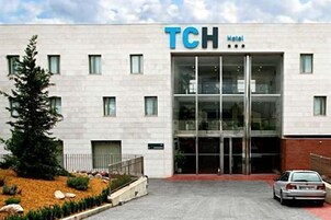 Edifício de um hotel TCH com fachada moderna, ampla entrada de vidro e área de estacionamento com um carro.