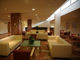 Um hall amplo com piso de mármore, várias áreas de estar incluindo sofás e cadeiras, uma mesa de recepção e arte abstrata nas paredes.