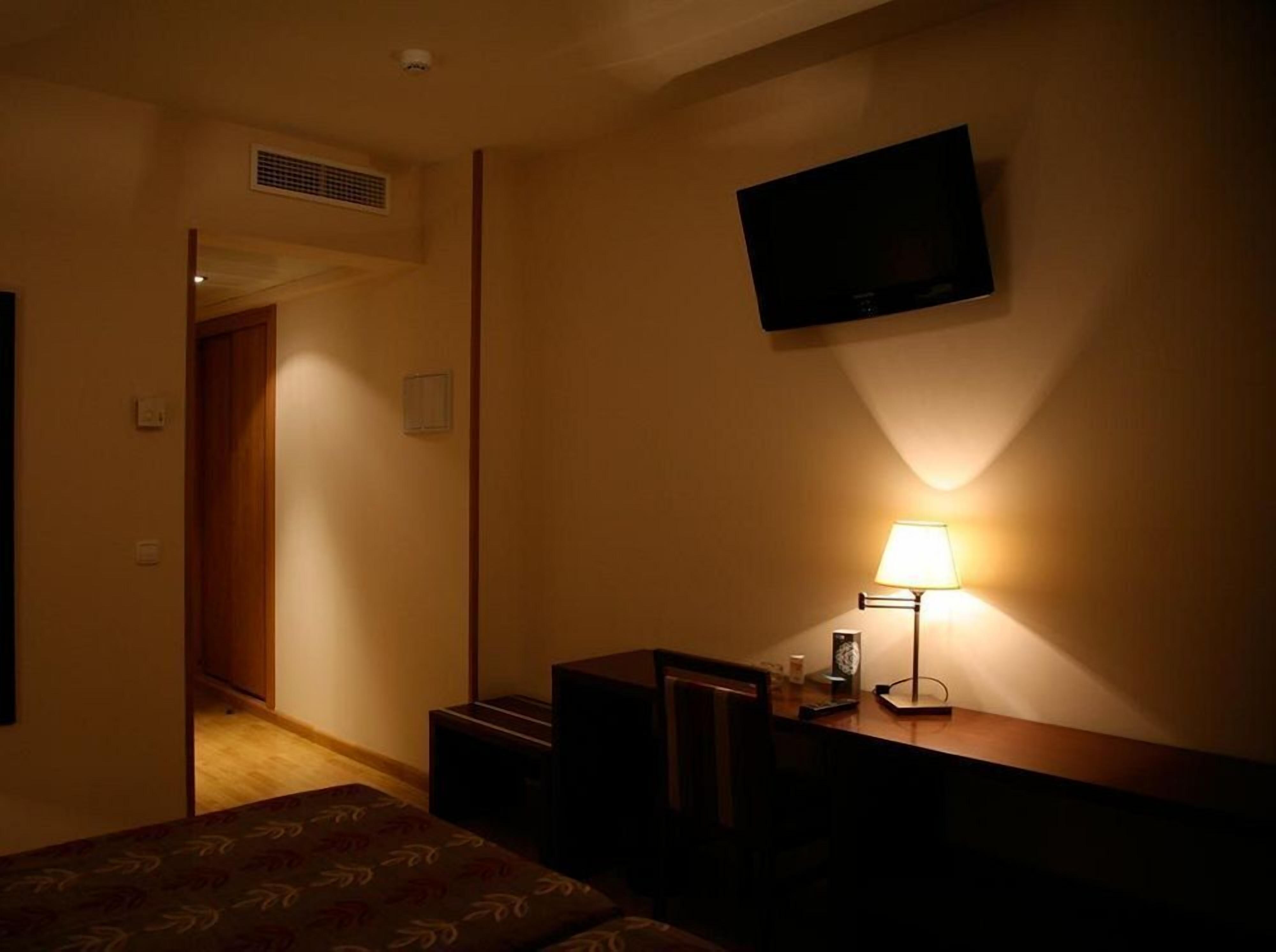 Habitación de hotel con cama, un escritorio con lámpara, televisor montado en la pared y una puerta que da a otra habitación.