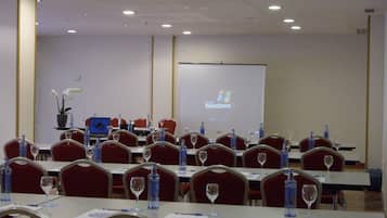 A conference room with rows of tables, each with a laptop, water bottle, and wine glass. A projector screen displays a presentation on a wall.