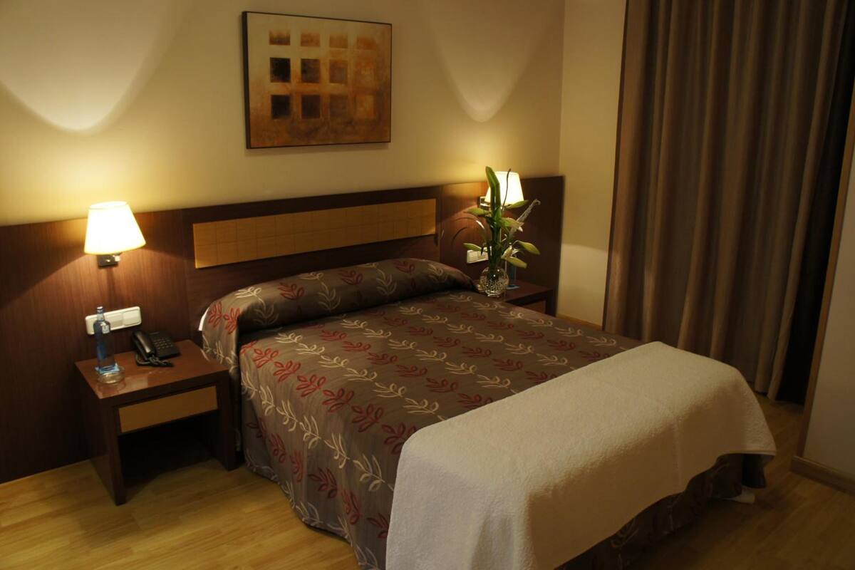 Habitación de hotel con cama, dos mesitas de noche, lámpara, un florero con flores y un cuadro en la pared.