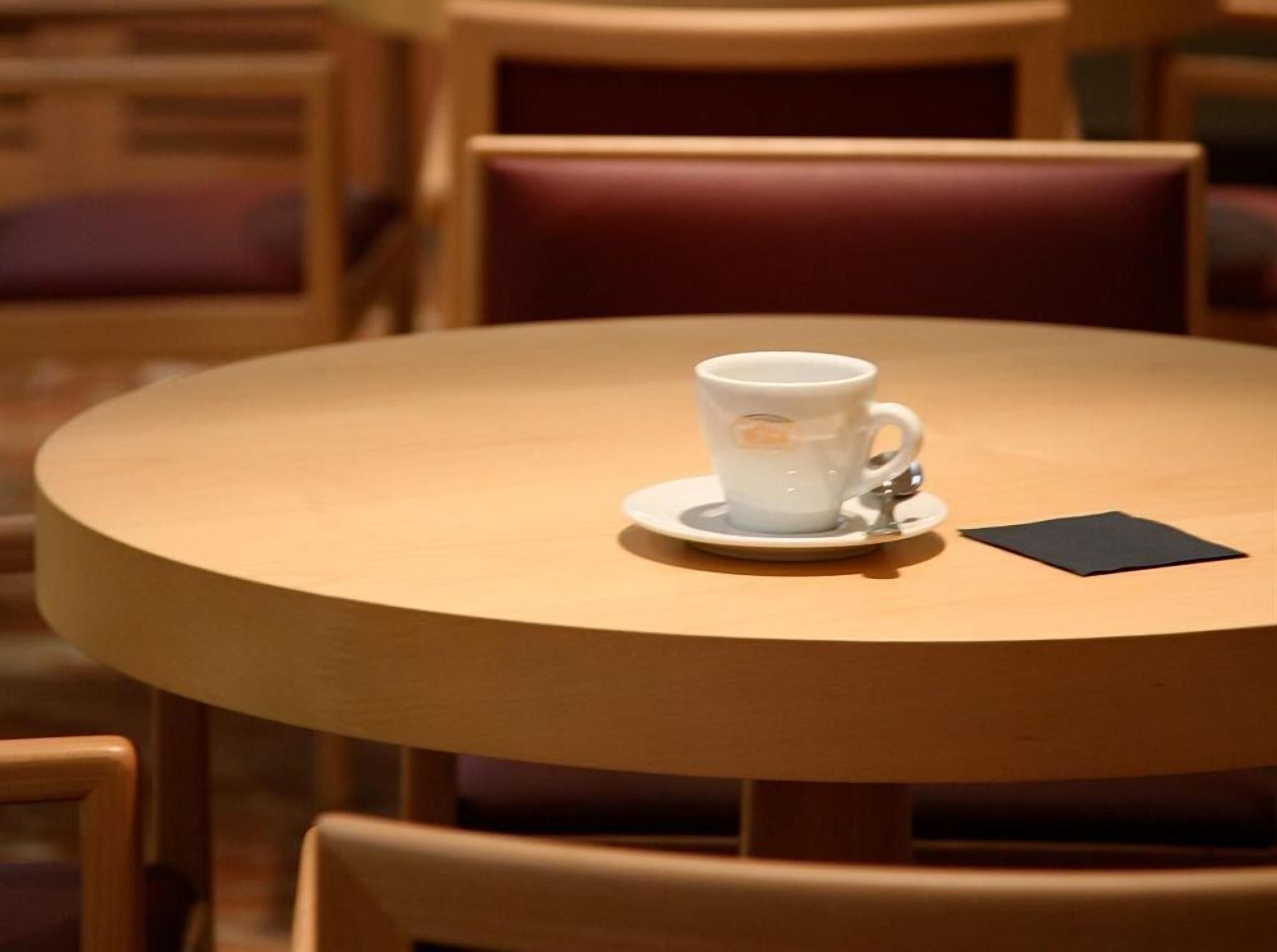 A round wooden table with a white ceramic cup and saucer, a black napkin, and a wooden chair.