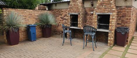 A brick outdoor kitchen with two barbecue grills, a seating area with two chairs, and potted plants.