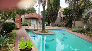 A swimming pool with a tiled deck, surrounded by greenery and a brick wall. - Rusplek Guesthouse and Conference Centre (Bloemfontein)
