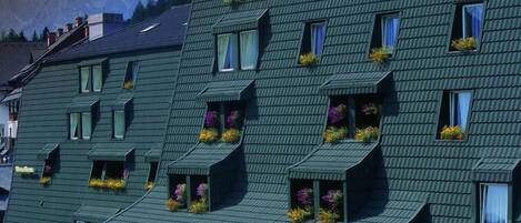 Un hotel llamado "Hotel Alpena" con un tejado pronunciado, varios balcones con jardineras y un paisaje montañoso de fondo.