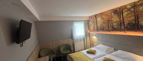 A hotel room with a bed, two chairs, a desk, a flat-screen TV, and a painting of a forest.