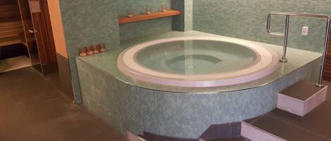 Una bañera de hidromasaje circular con recinto de vidrio, rodeada de suelo de baldosas y un estante de madera con velas.