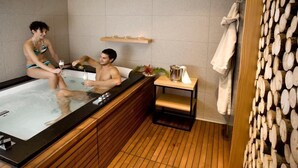 Habitación con piso de madera, una bañera de hidromasaje, una mesita con una botella y una planta, y un perchero para toallas.