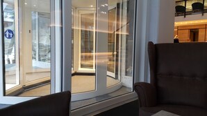 Un vestíbulo de hotel con pared de vidrio, una zona de estar con sillas y una entrada visible.