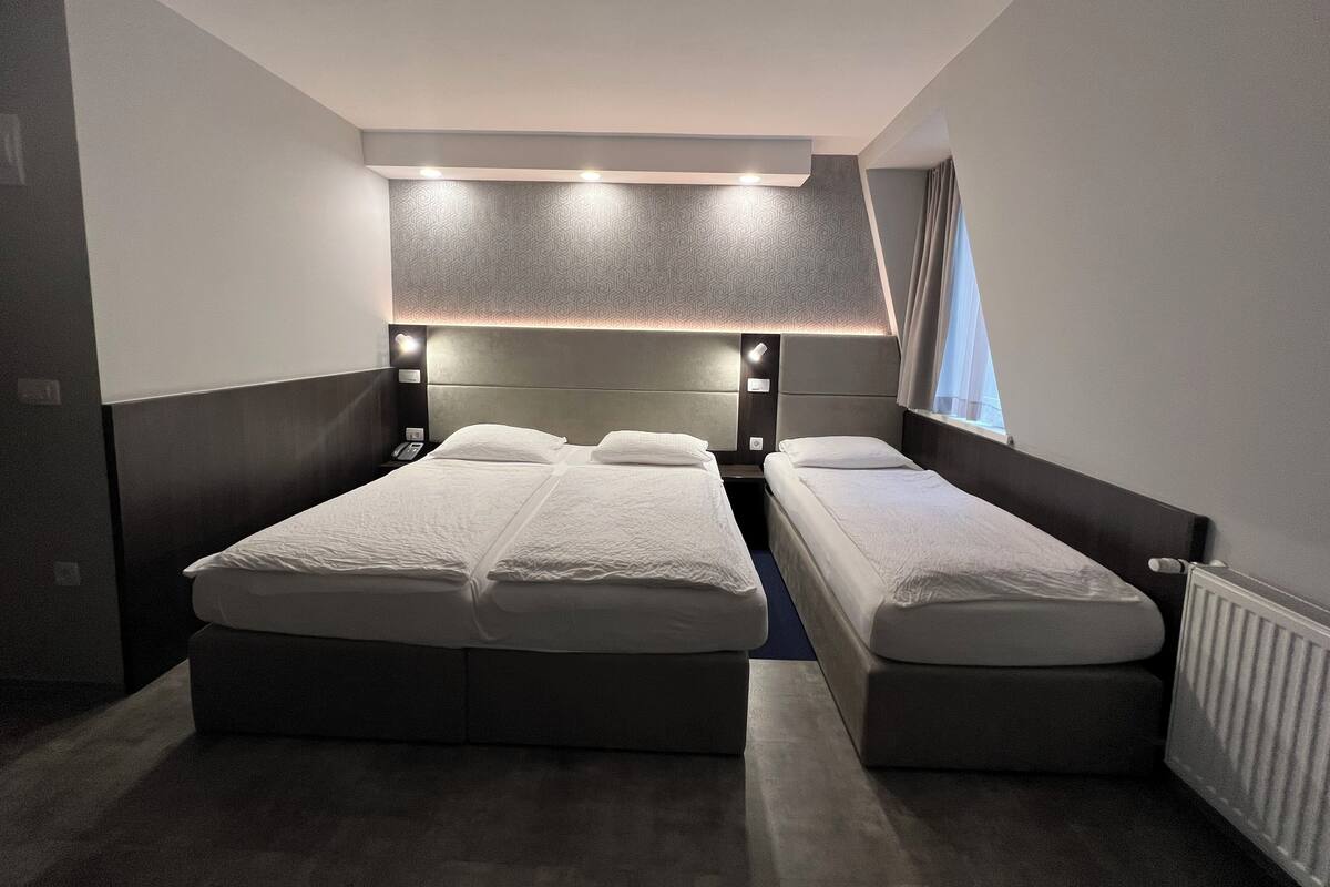 A hotel room with two beds, a headboard with built-in lighting, a window with curtains, and a radiator.
