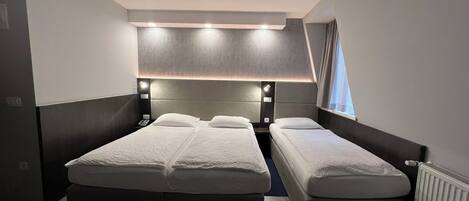 A hotel room with two beds, a headboard with built-in lighting, a window with curtains, and a radiator.