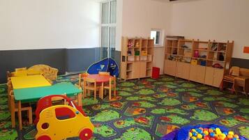 A children's playroom with a colorful carpet, play tables, chairs, and a ball pit.