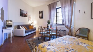 A bedroom with a bed, a desk, a sofa, a dining table with chairs, and a window with curtains. - 1900 Artevita (Florence)
