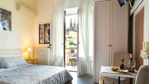 A hotel room with a bed, a table with a plate of food, a bottle of wine, and a chair. There is a window with curtains, a radiator, and a wall-mounted TV. - 1900 Artevita (Florence)