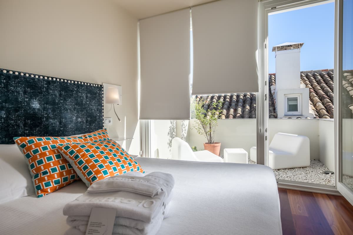 Habitación de hotel con cama, un ventanal grande, vistas de la ciudad y un balcón con mobiliario exterior.