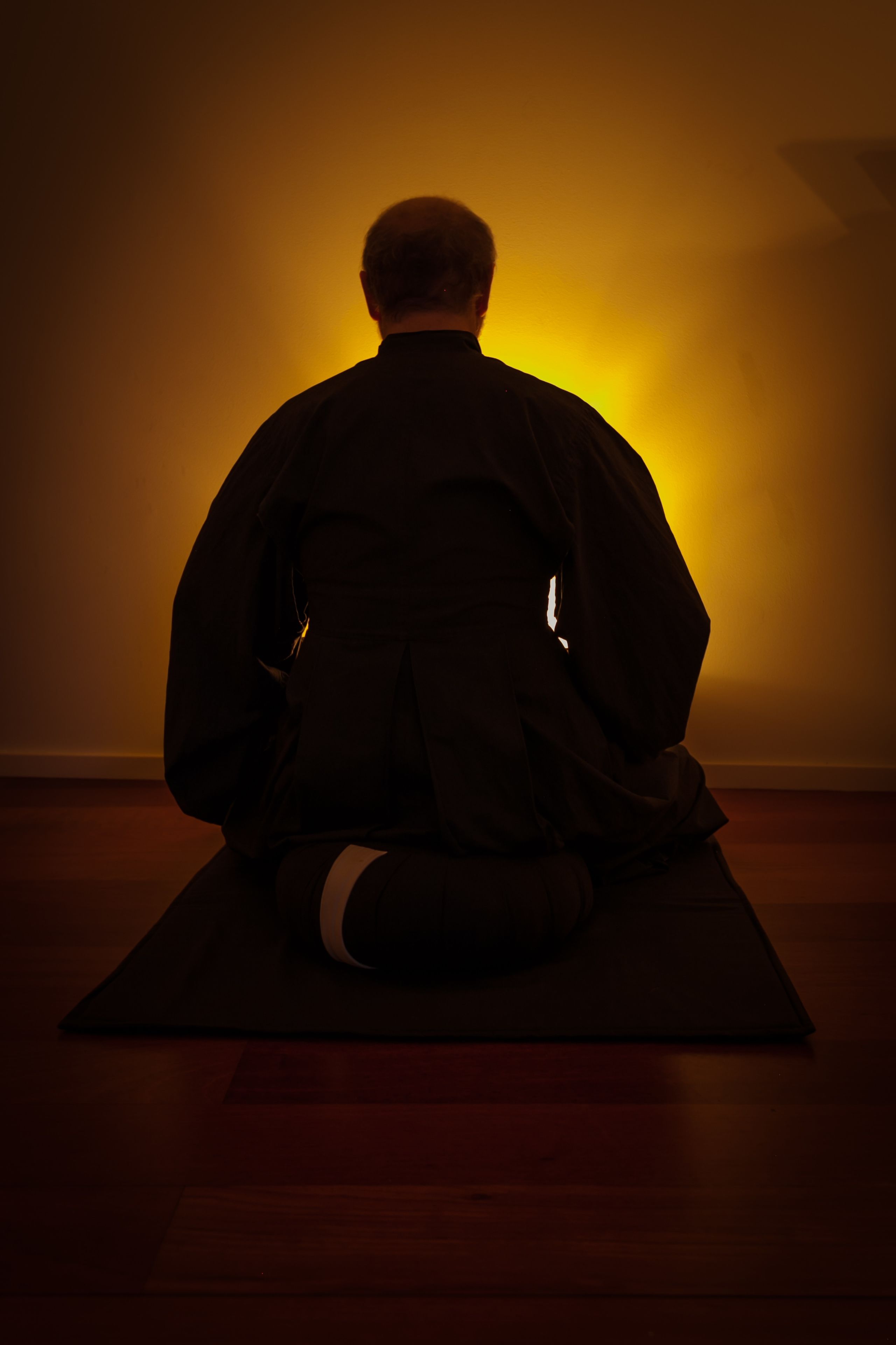 Una persona sentada en un tapete en una habitación tenuemente iluminada, con una pared lisa y piso de madera.