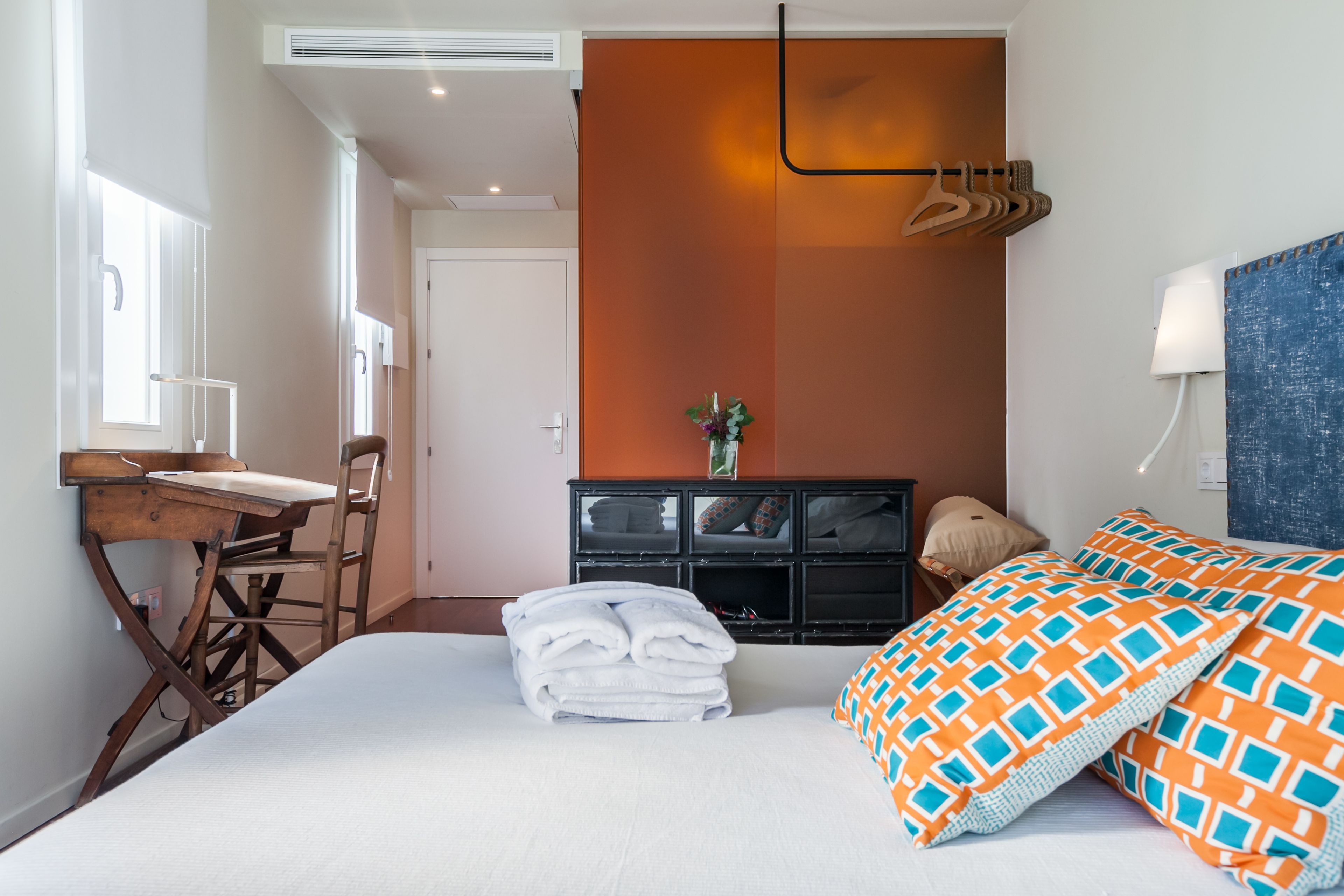 A hotel room with a bed, a desk, a chair, a dresser, and a hanging coat rack.