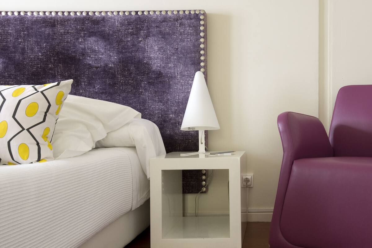 Una cama bien hecha con un cojín estampado, una lámpara de noche blanca y una silla morada en una habitación con cabecera texturizada.