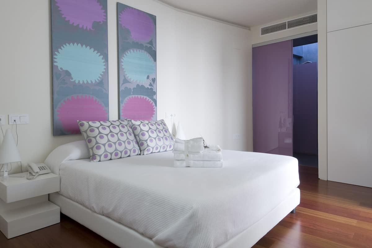 Un dormitorio moderno con una cama bien hecha, una mesita de noche con lámpara y un cuadro abstracto grande en la pared.