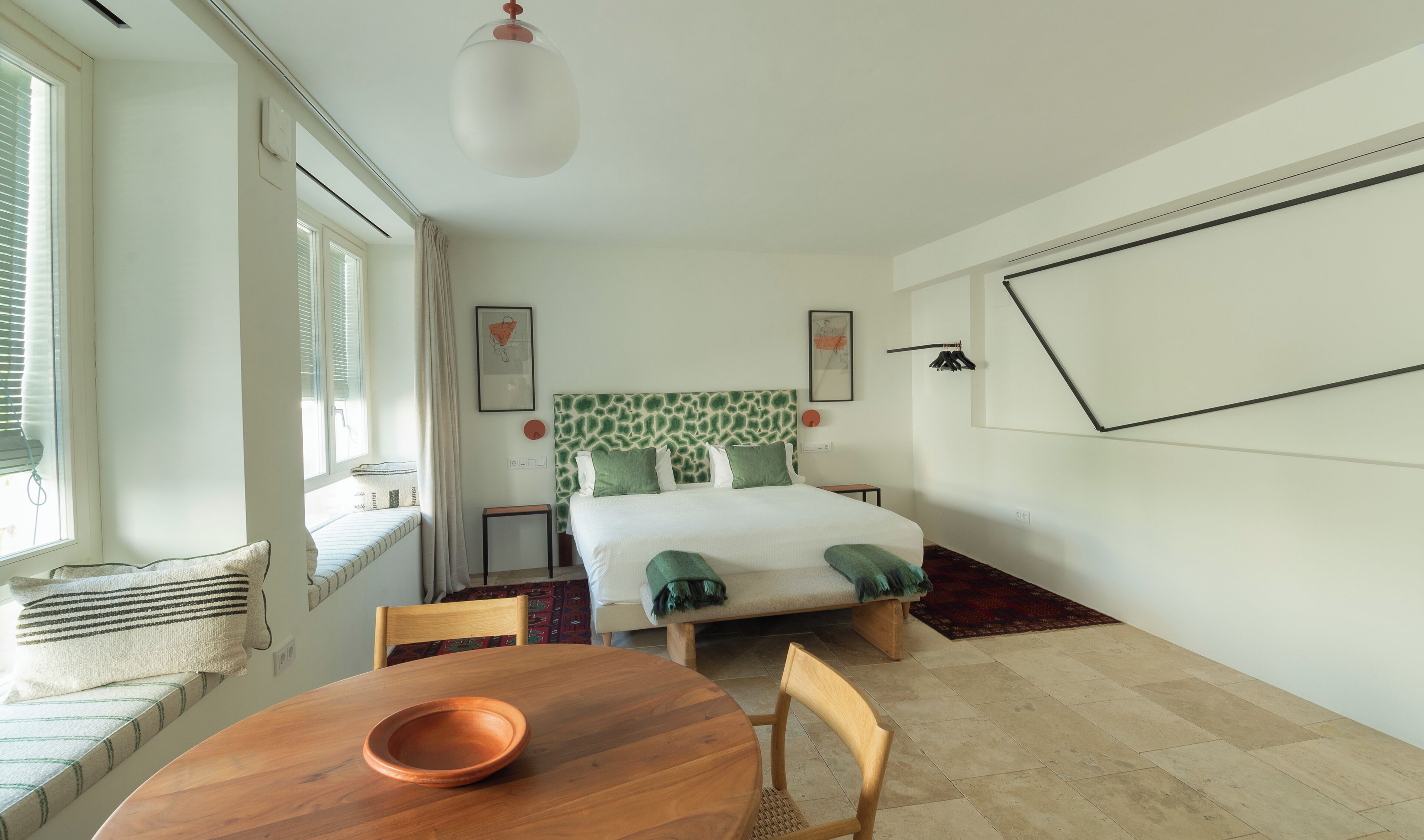 Un dormitorio con cama, una ventana con persianas, una mesita de noche, una silla y una mesa de comedor con un bol.