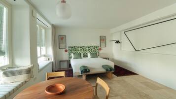 Un dormitorio con cama, una ventana con persianas, una mesita de noche, una silla y una mesa de comedor con un bol.