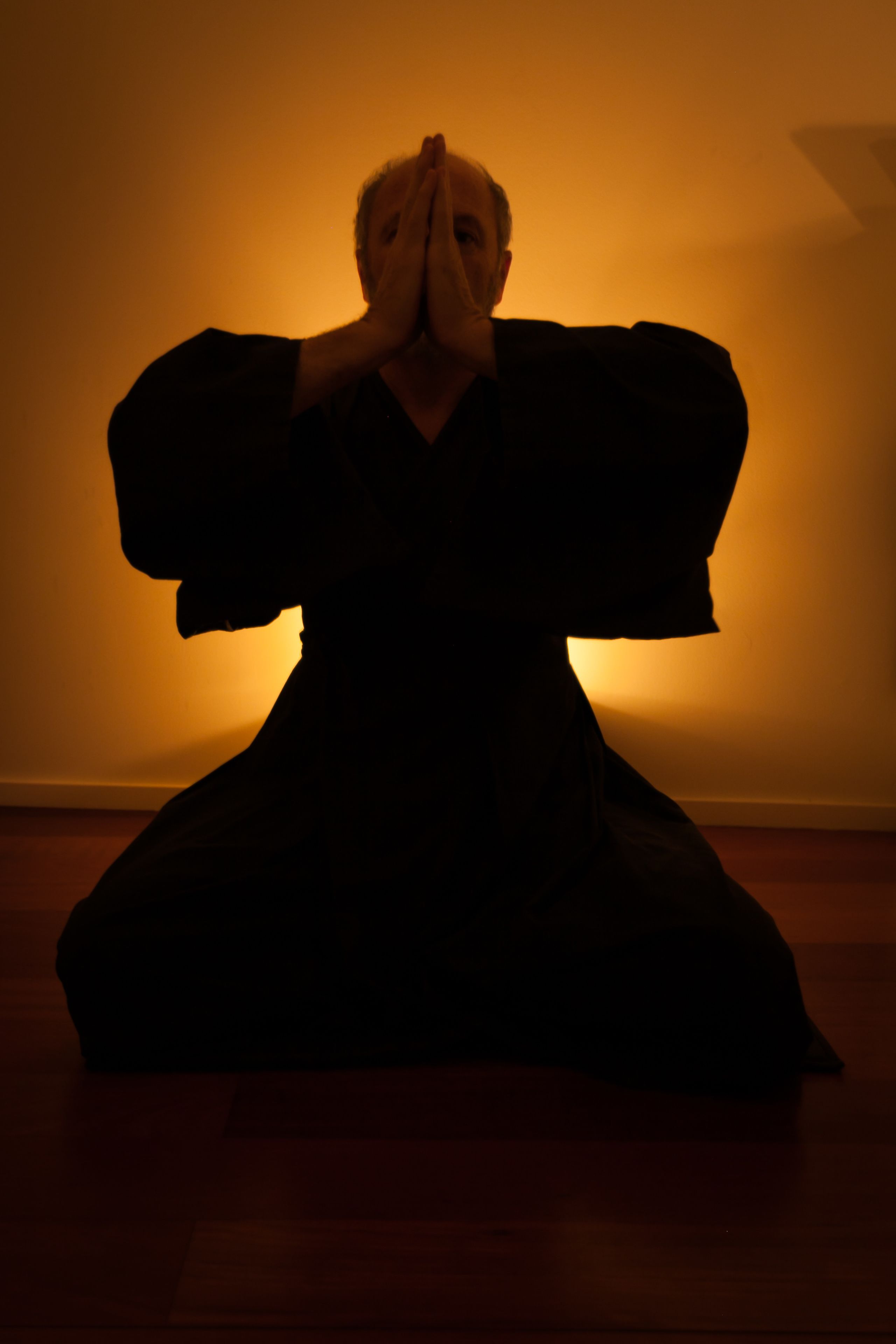 A person in a dark outfit sitting on a wooden floor with a plain wall in the background.