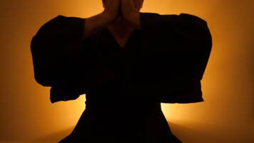 A person in a dark outfit sitting on a wooden floor with a plain wall in the background.