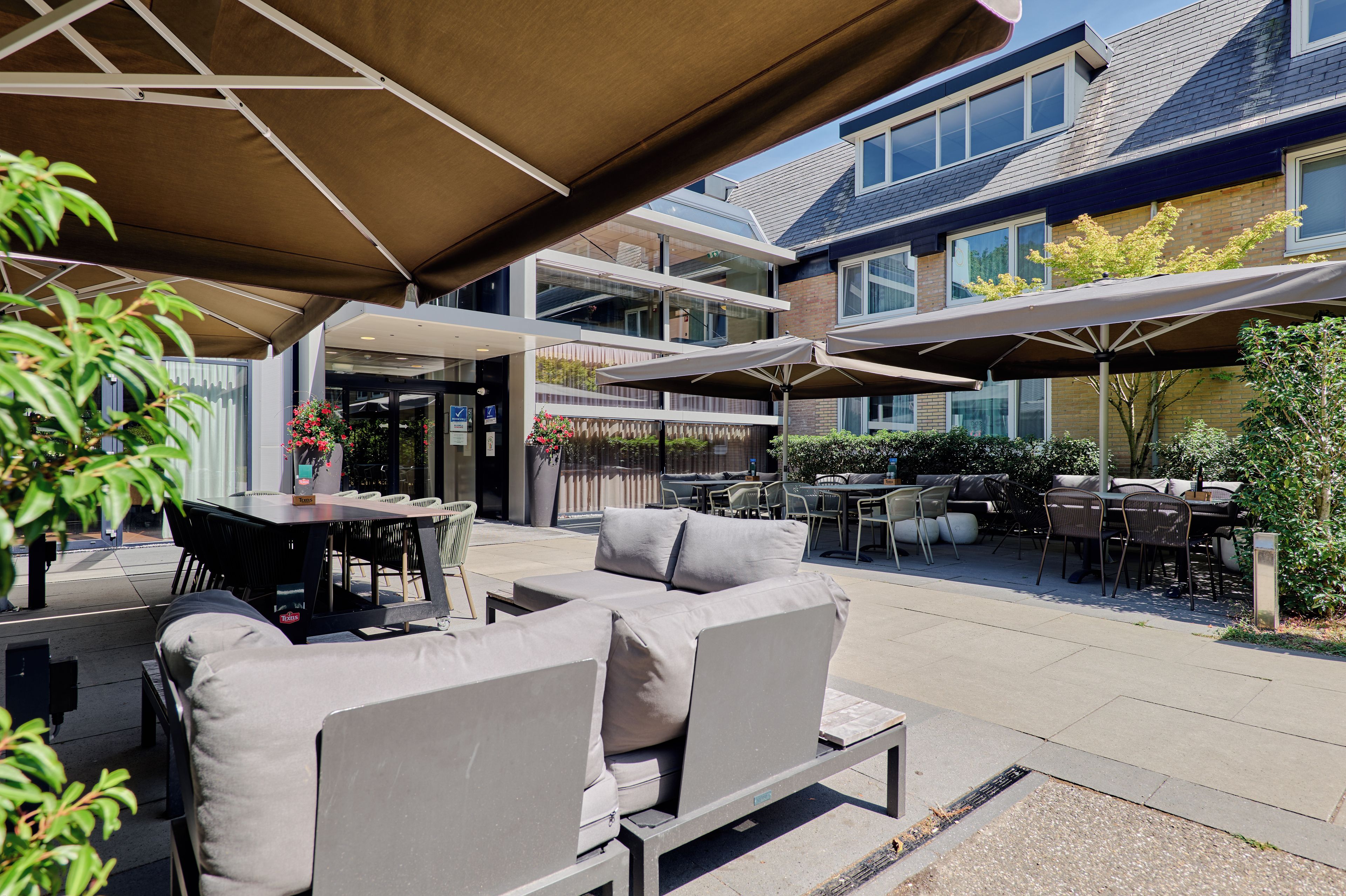 An outdoor patio area with multiple tables and chairs, some under umbrellas, and a building with large windows in the background.