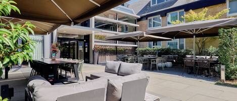 An outdoor patio area with multiple tables and chairs, some under umbrellas, and a building with large windows in the background.