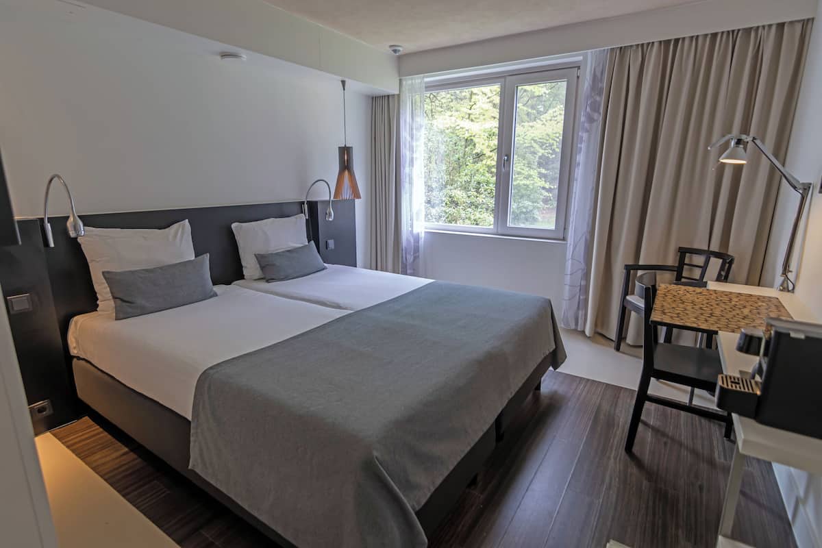 A modern hotel room with a large bed, bedside lamps, a desk with a chair, and a window with curtains.