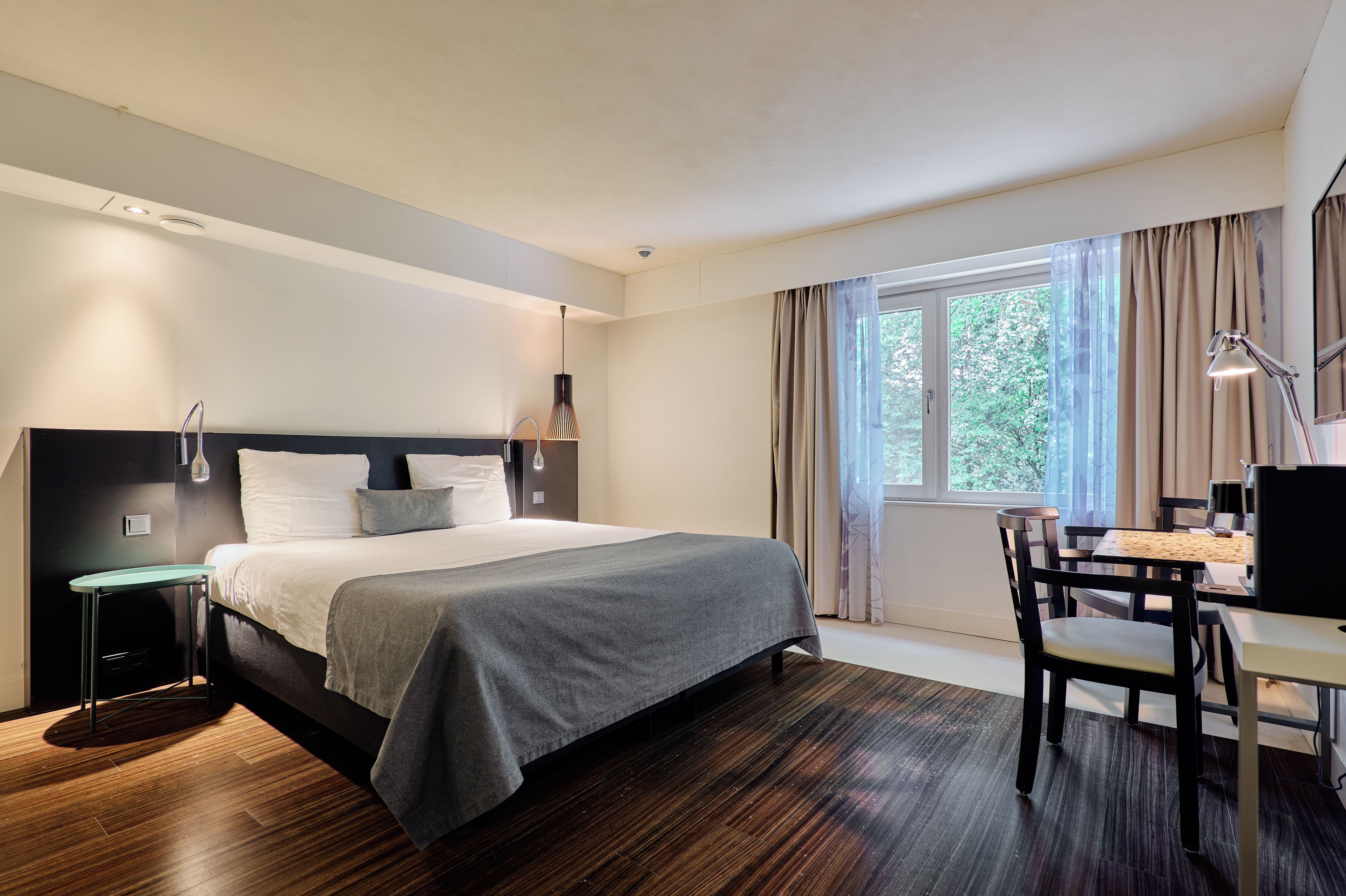 A modern bedroom with a bed, bedside table, desk, chair, and a window with curtains.