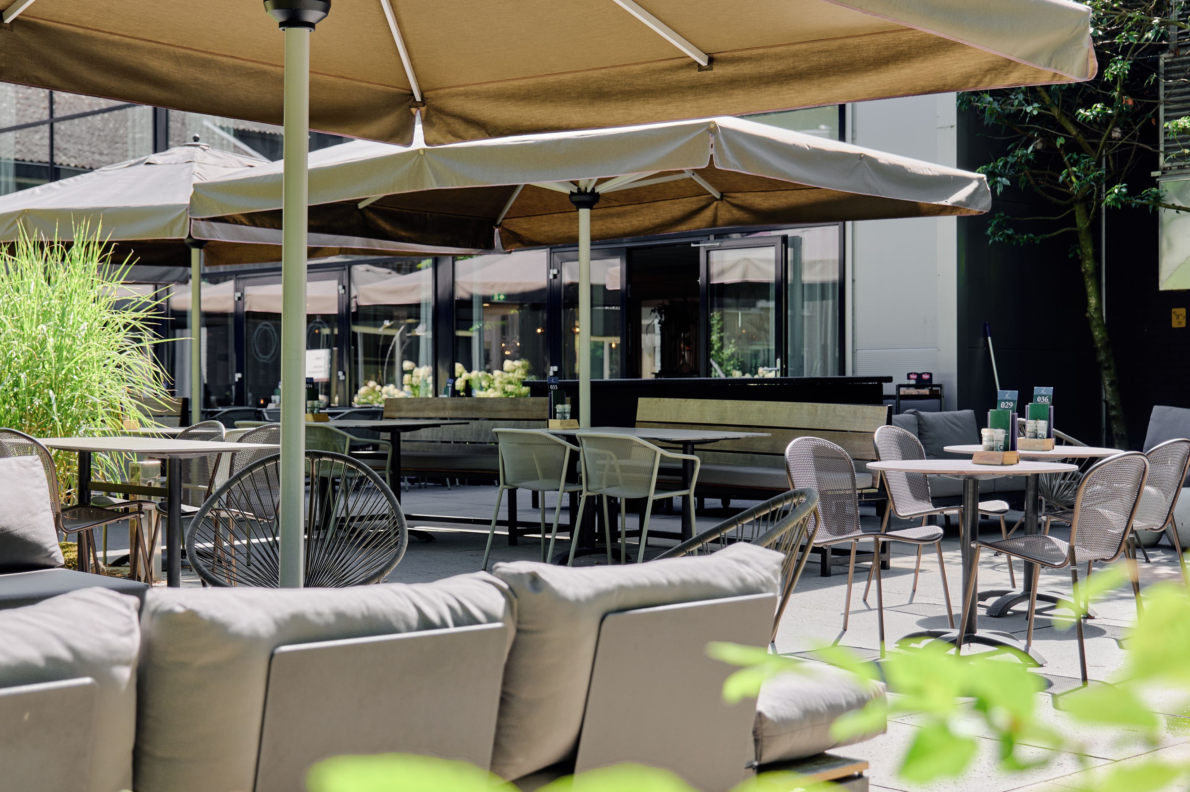 An outdoor seating area with tables and chairs, some covered by umbrellas, and a view of the interior through glass walls.