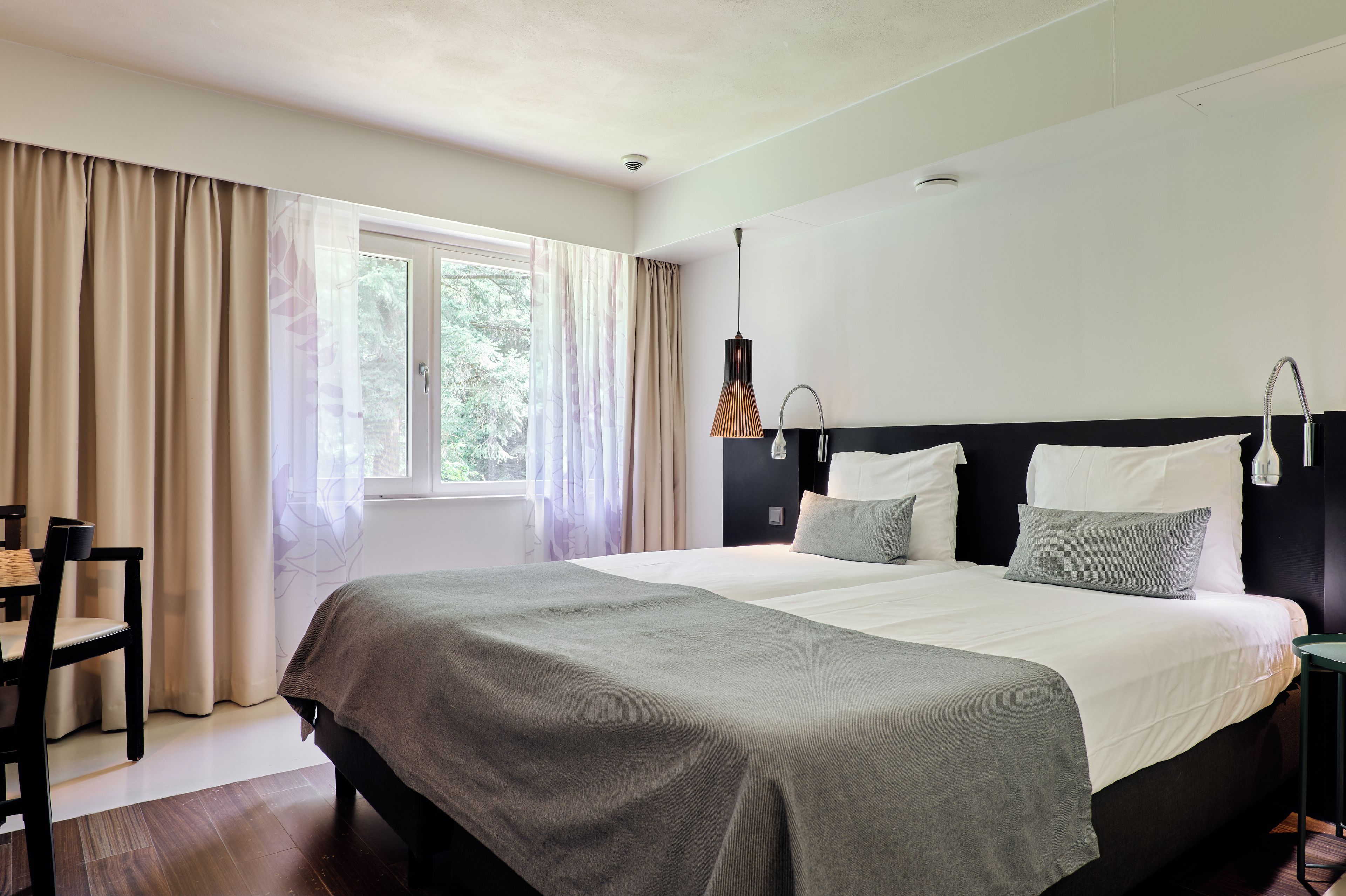 A hotel room with a bed, a chair, a window with curtains, and a nightstand.