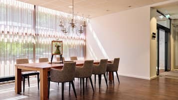Un salón de conferencias con una mesa de madera, sillas estampadas, una lámpara de araña y ventanas grandes con cortinas translúcidas.