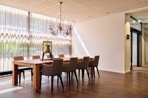 Un salón de conferencias con una mesa de madera, sillas estampadas, una lámpara de araña y ventanas grandes con cortinas translúcidas.