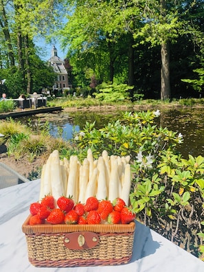 Un panier en osier contenant des asperges et des fraises est posé sur une table près d’un plan d’eau. On aperçoit des arbres et un bâtiment au loin.