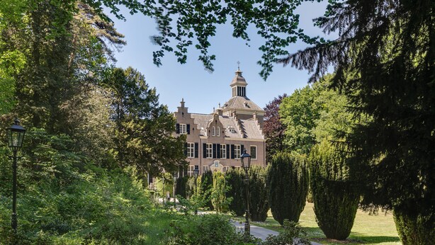 Un edificio grande e histórico con una torre destacada, rodeado de vegetación exuberante y un jardín bien cuidado.