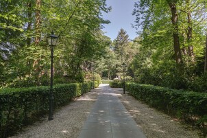 Um caminho ladeado por sebes e árvores bem aparadas, conduzindo a uma construção distante.
