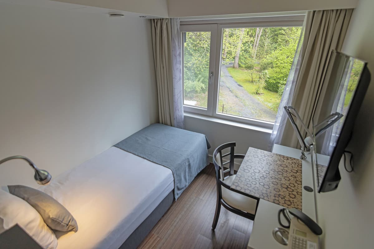 A small, well-lit room with a single bed, a desk with a chair, and a window offering a view of greenery outside.