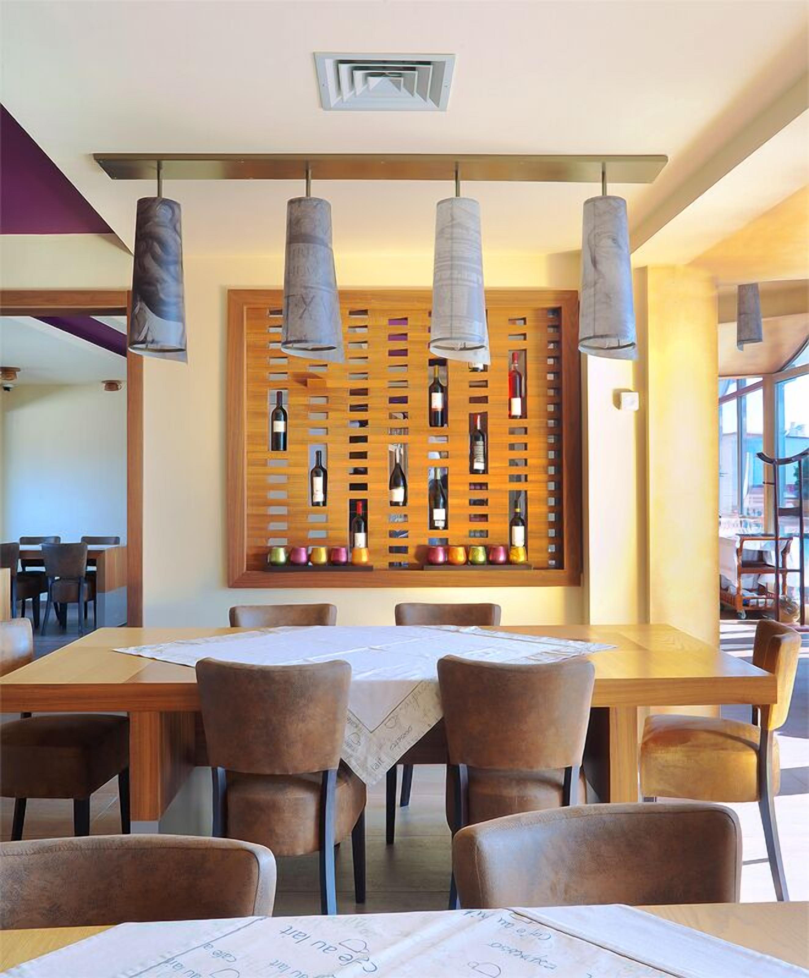A restaurant with a wooden wine rack displaying various bottles, hanging pendant lights, and a long wooden table with chairs.