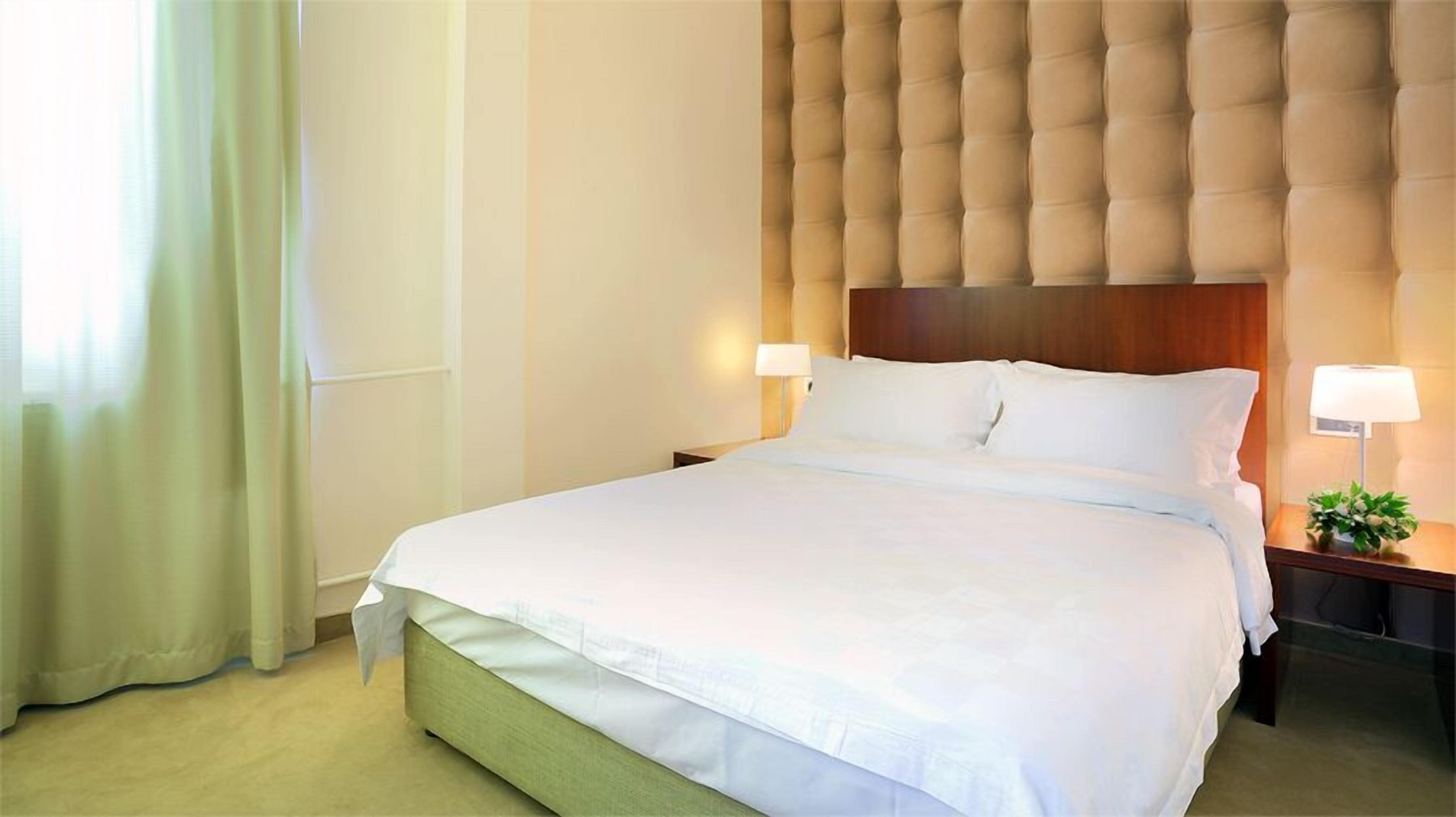 A hotel room with a large bed, two bedside tables with lamps, a tufted headboard, and a window with curtains.