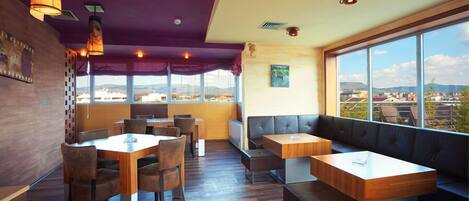 Un restaurante con mesas y bancos de madera, grandes ventanales que ofrecen vistas a la ciudad y un techo morado.