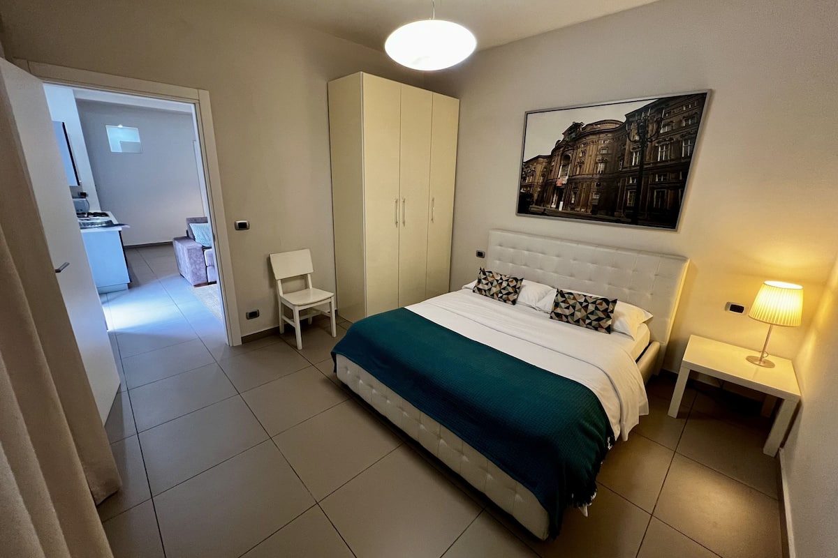 Een slaapkamer met een bed, een stoel, een kledingkast, een nachtkastje en een schilderij aan de muur.